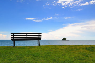 Empty bench on grassy field by sea against blue sky