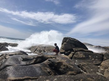 Man crouching on rock at beach against sky