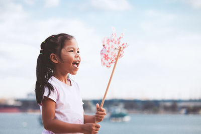 Side view of cheerful girl holding pinwheel toy at beach against sky