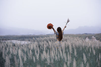 Rear view of woman with arms raised standing amidst plants on field against sky