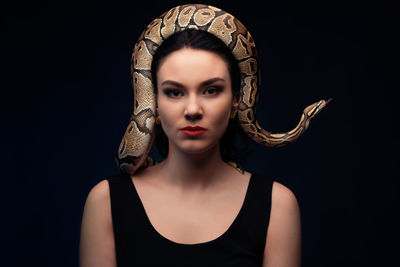 Portrait of young woman with snake on head against black background