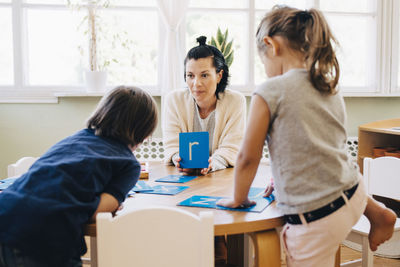 Confident female teacher holding letter r in front of students at table in classroom