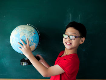 Portrait of boy wearing eyeglasses while holding globe against black board