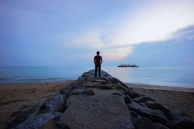 Rear view of man standing on rock at beach against cloudy sky during sunset