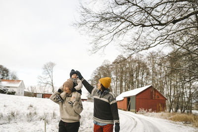 Mature woman adjusting knit hat of daughter sitting on girlfriend's shoulder