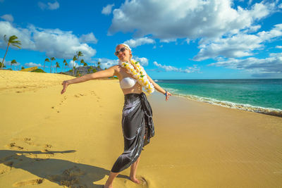 Mature woman wearing floral garland while standing with arms outstretched at beach against sky