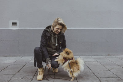 Smiling blond woman crouching while playing with pomeranian dog against wall