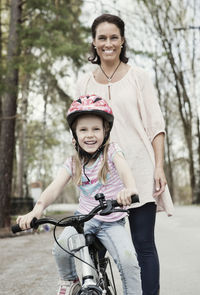 Portrait of happy girl riding bicycle while mother standing behind her on road