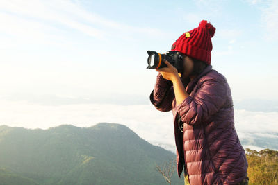 Side view of woman wearing jacket photographing with digital camera against mountains