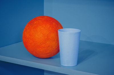 Orange papier-mache ball and plastic cup on the blue shelf - geometric design with space for copy.