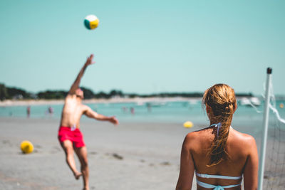 Couple playing volley ball while standing at beach against clear blue sky during sunny day