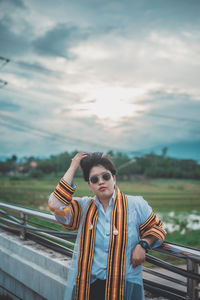 Portrait of young woman standing by railing against sky