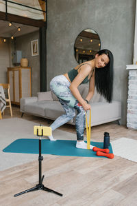 Young woman in sportswear does an exercise with an elastic band and uses a smartphone on a tripod