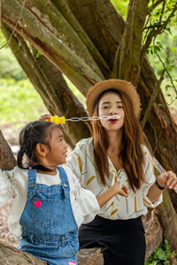 Portrait of happy woman blowing bubble with daughter while sitting on tree