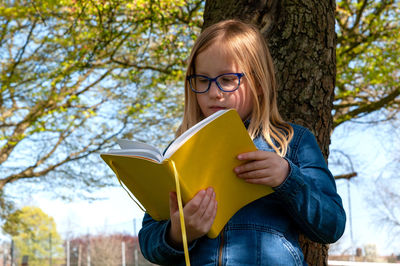 Young school age girl reading from yellow textbook in park.