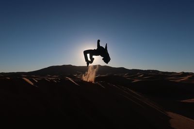 Silhouette person backflipping at desert against clear blue sky during sunset