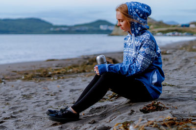 Young woman holding insulated drink container while sitting at beach