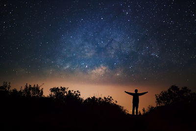 Silhouette man with arms outstretched standing amidst plants against star field at night