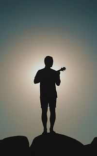 Silhouette mid adult man playing guitar while standing on rock formation against blue sky