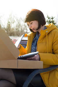 A happy brunette woman in a yellow jacket on the street eagerly opens a new laptop.