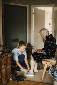 Female nurse helping senior woman to wear shoe in living room at home