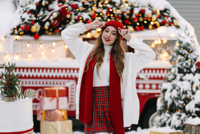 A beautiful young woman with red lips in a warm hat stands by decorated christmas van on the street