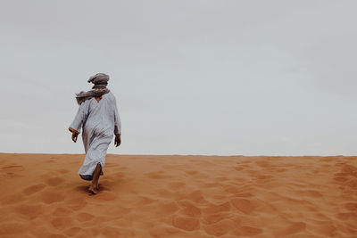 Rear view of man walking on sand dune against clear sky