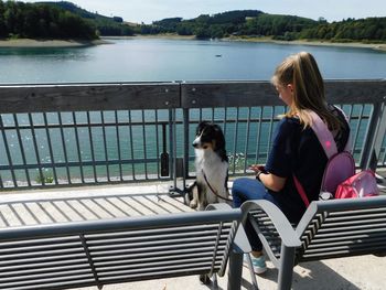 Rear view of girl with dog sitting on bench against lake