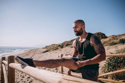 Man stretching on wooden railing at beach against sky