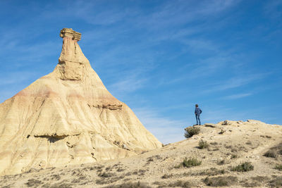 Rear view of a man with hat and sunglasses standing on desert dunes against a rock peak