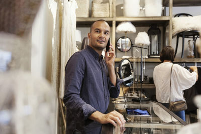 Smiling male owner using phone while customer looking at clothes in store