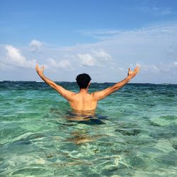 Rear view of shirtless man with arms outstretched standing in sea against sky