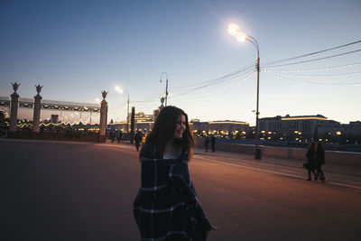 Happy woman wrapped in blanket standing on road against clear sky at dusk