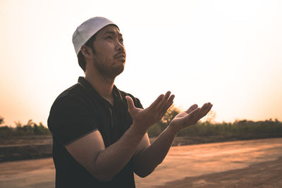 Side view of young man holding hat against sky during sunset