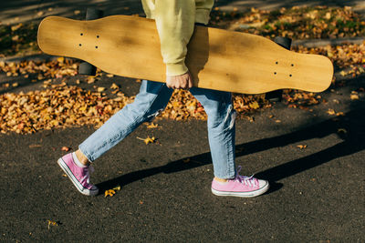 Girl holding a longboard while walking down the street with fall leaves in the background