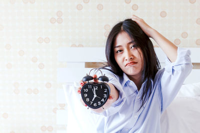 Portrait of beautiful woman showing alarm clock while sitting on bed