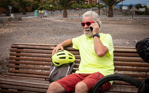 Senior man answering smart phone while sitting on bench with cycling helmet