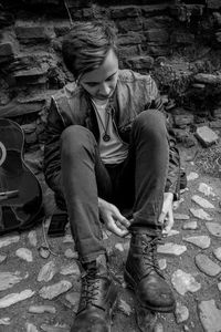 High angle view of young man adjusting jeans while sitting on cobbled street by guitar