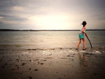 Rear view of girl holding stick standing at beach against sky