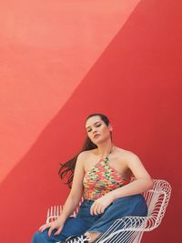 Portrait of beautiful young woman sitting on chair against red wall