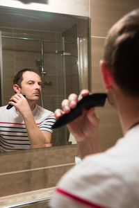 Young man reflecting on mirror while trimming beard in bathroom