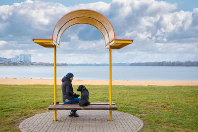 Rear view of woman sitting on bench with dog against sky