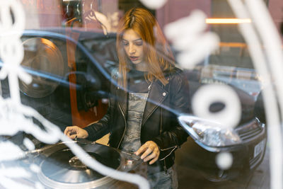 Through window of woman playing vintage record player while standing in vinyl store located on street near road with car