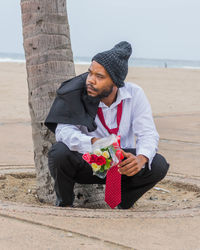 Full length of man in formalwear holding bouquet while crouching at sandy beach