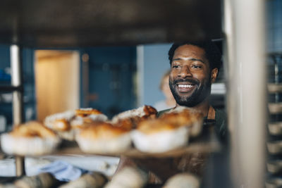 Smiling professional male baker arranging fresh buns in cooling rack at bakery