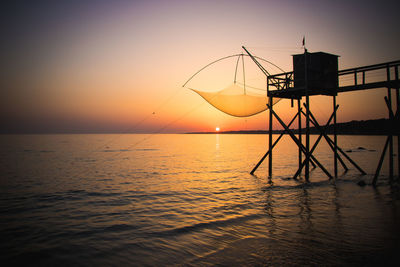 Silhouette fishing net on sea against sky during sunset