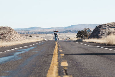 Man carrying skateboard while standing on road against clear sky