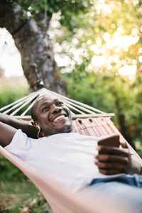 Smiling mid adult man listening music through mobile phone while relaxing on hammock in backyard