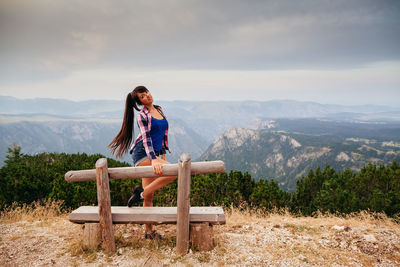Portrait of woman standing on mountain by bench against sky