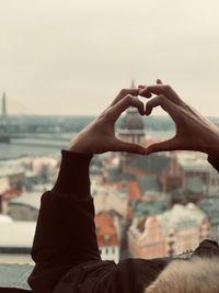 Cropped hands of person making heart shape against buildings and sky